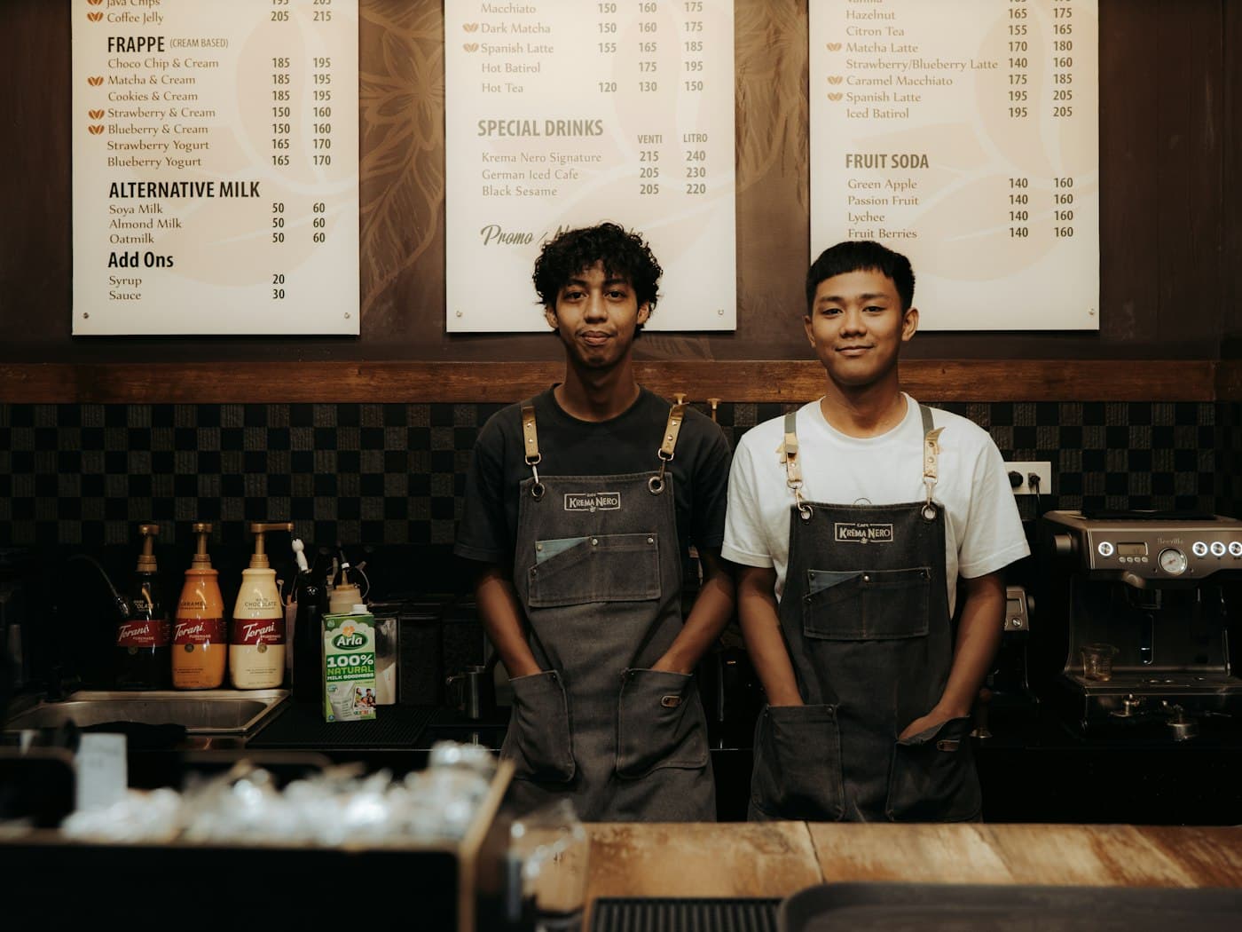 Dois baristas sorrindo atrás do balcão de uma cafeteria